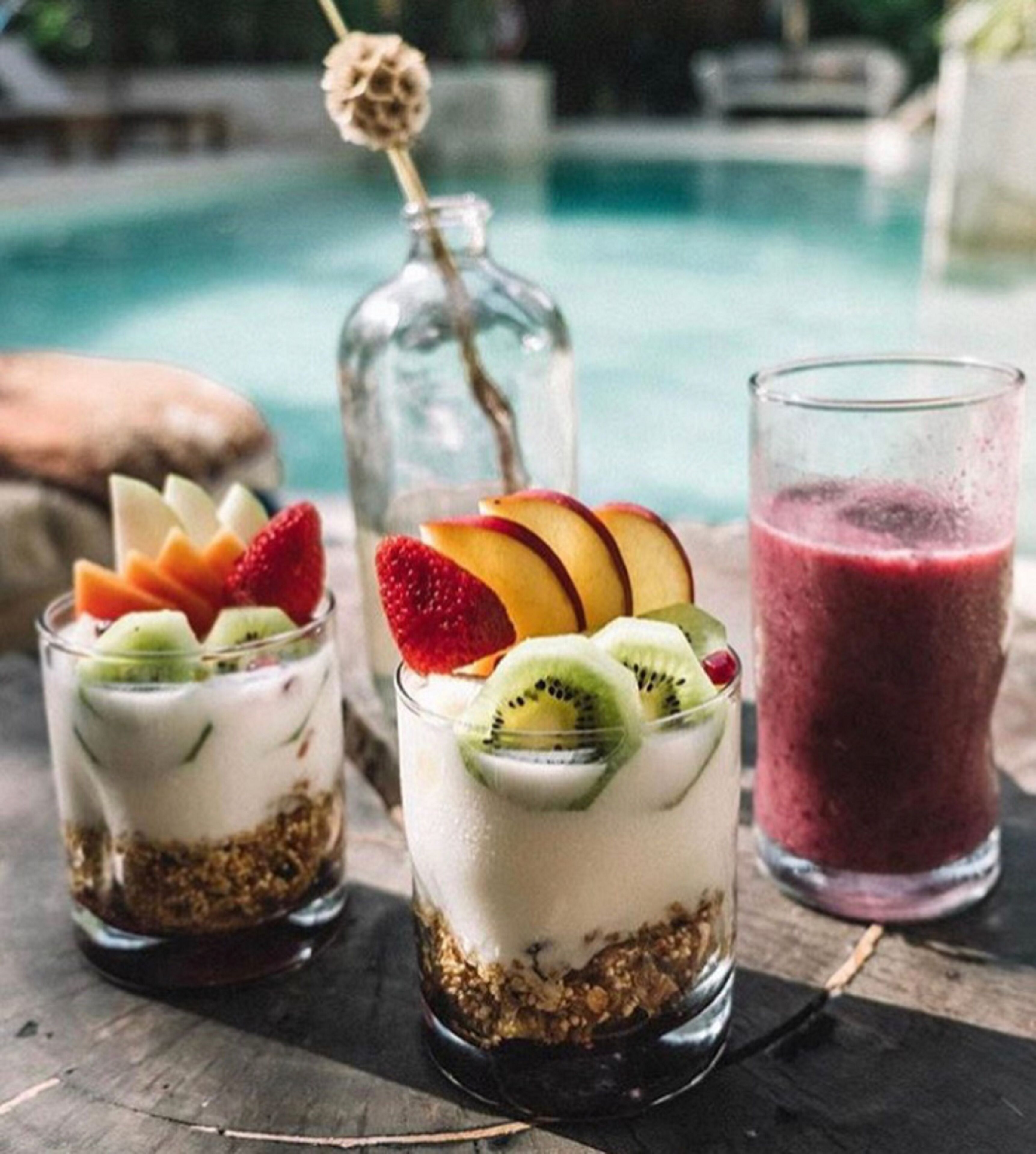 Tres vasos de parfait de frutas y un batido sobre una mesa de madera junto a una piscina.