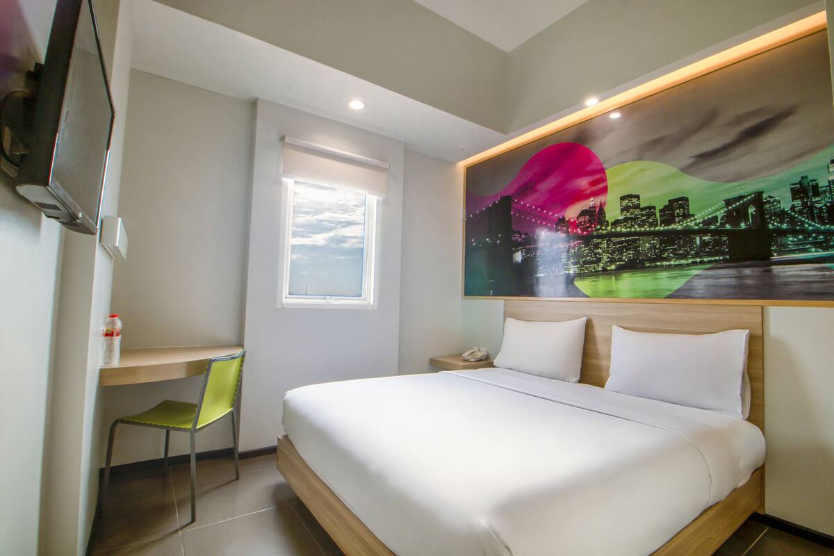A hotel room with a bed, a desk, a chair, a TV, and a cityscape painting on the wall.