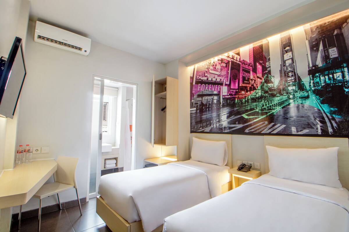 A hotel room with two beds, a desk, a chair, and a large cityscape artwork on the wall.