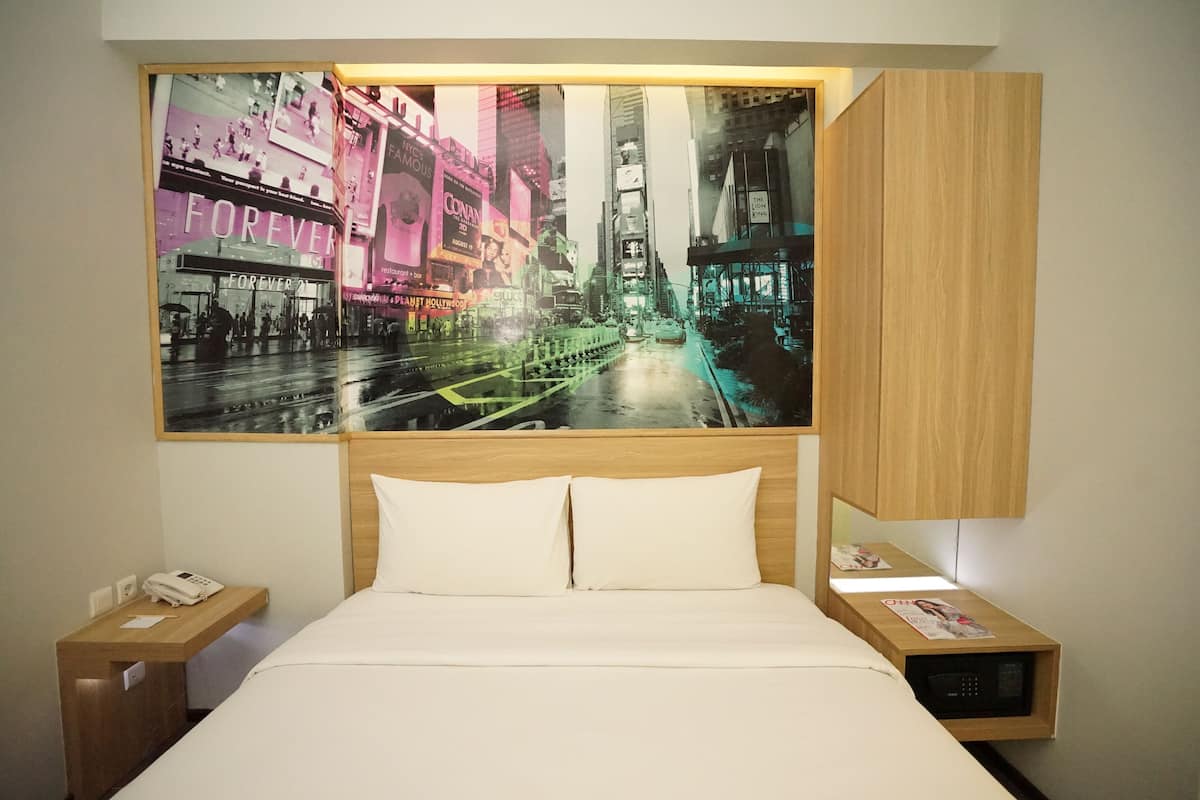 A hotel room with a bed, a nightstand, a bedside table, a phone, a magazine, and a large cityscape painting on the wall.