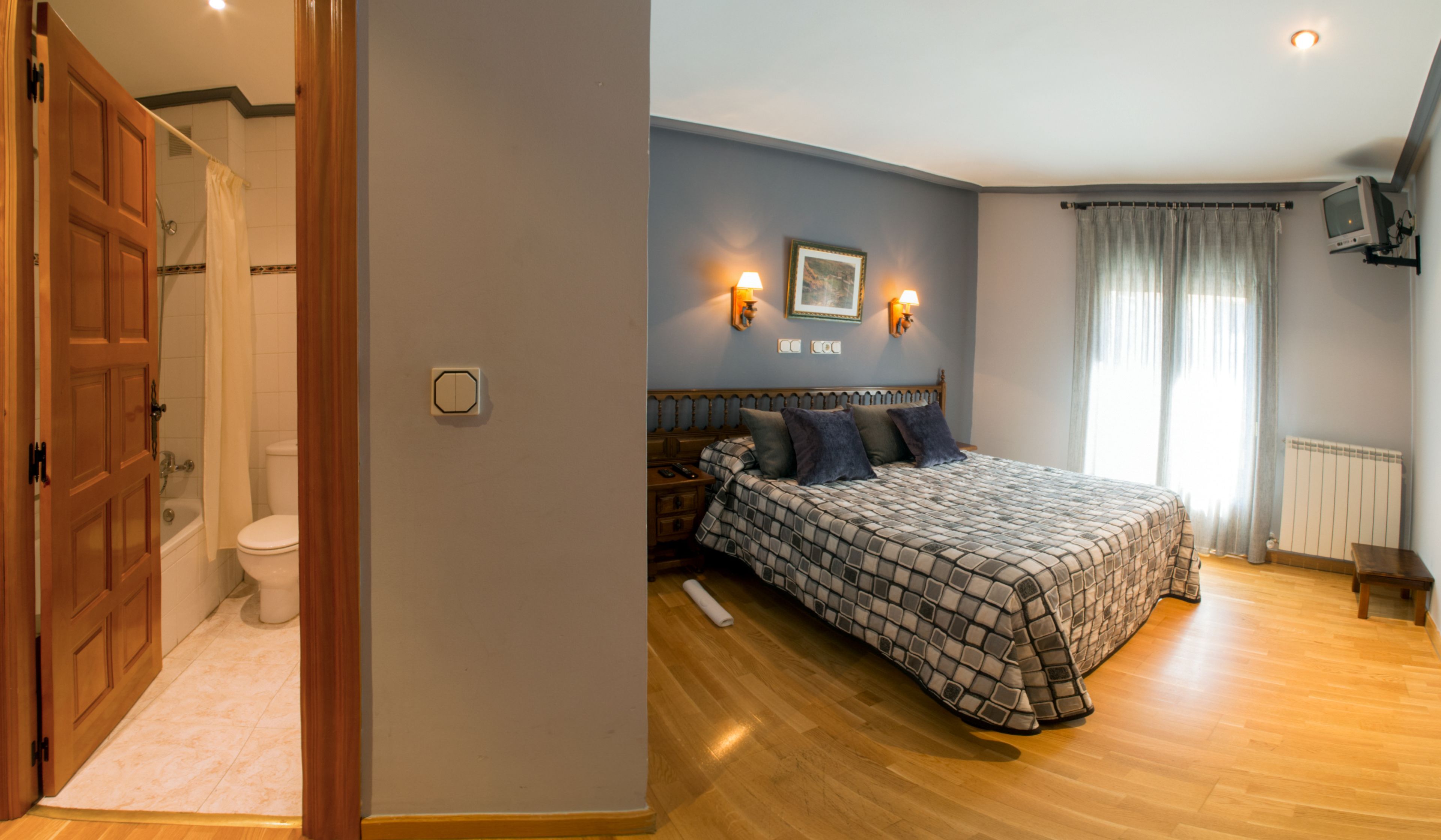 Ein Schlafzimmer mit Holzboden, einem Bett mit gemustertem Überwurf, einem Nachttisch, einem an der Wand montierten Fernseher und einem Fenster mit Vorhängen.