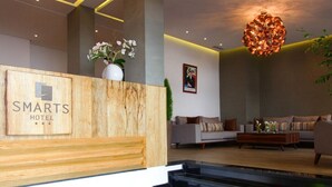 Uma área de recepção com balcão de madeira identificado como "SMARTS HOTEL", um lustre moderno e um espaço de estar com sofás e mesa de centro.