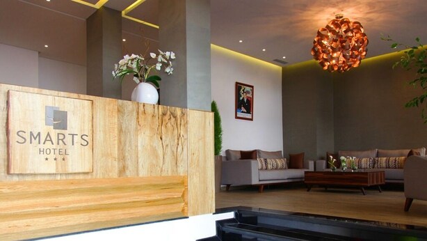 A reception area with a wooden reception desk labeled "SMARTS HOTEL," a modern chandelier, and a seating area with sofas and a coffee table. - Smarts Hotel (Rabat)