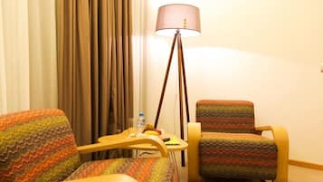 A hotel room with a wooden armchair, a cushioned armchair, a small table with a bottle and a banana, and a floor lamp.