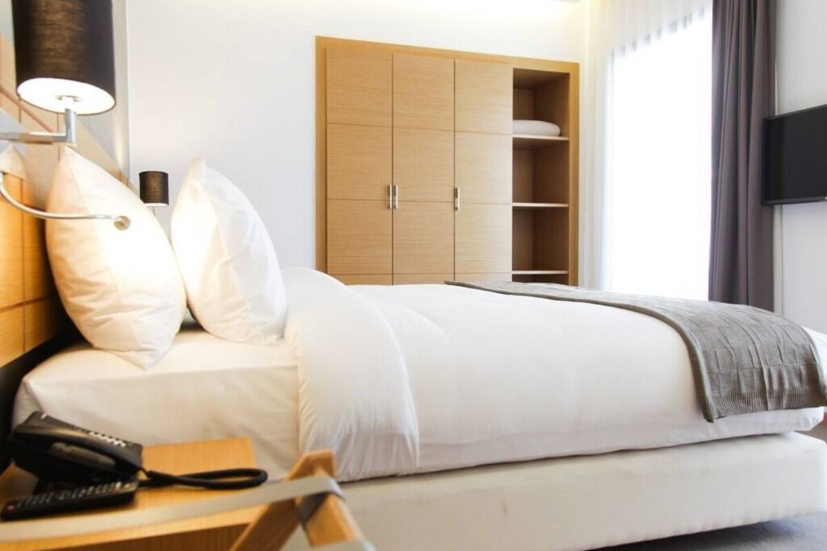 A modern hotel room with a large bed, a bedside table with a lamp, a wardrobe, and a TV mounted on the wall.