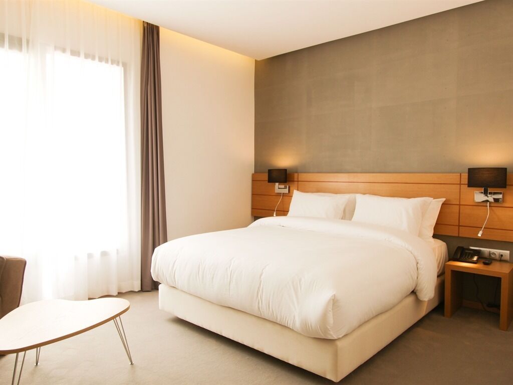 Ett hotellrum med en stor säng, sängbord, en stol och utsikt över omgivningarna genom tunna gardiner.
