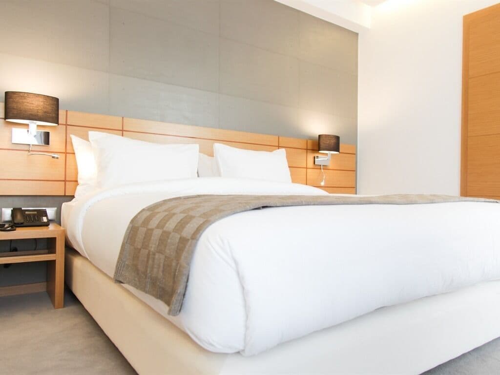 A modern hotel room with a large bed, wooden headboard, bedside tables, and wall-mounted lamps.