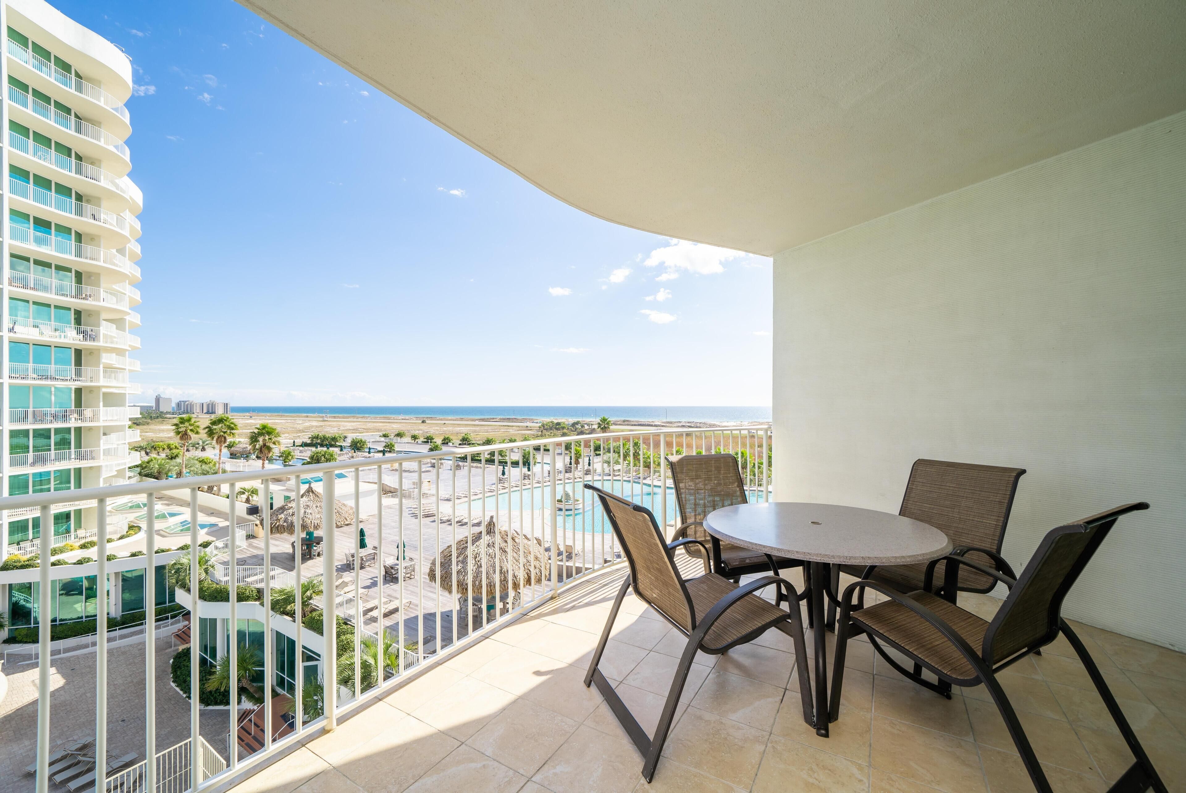 A balcony with a round table and chairs, overlooking a pool and a beach.