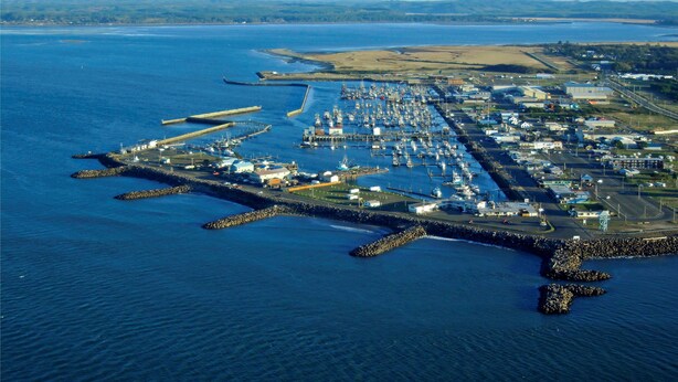 Vue aérienne d’un port de plaisance situé sur la côte, avec ses quais et ses bateaux, entouré d’une digue rocheuse et bordé par une ligne de rivage lointaine.