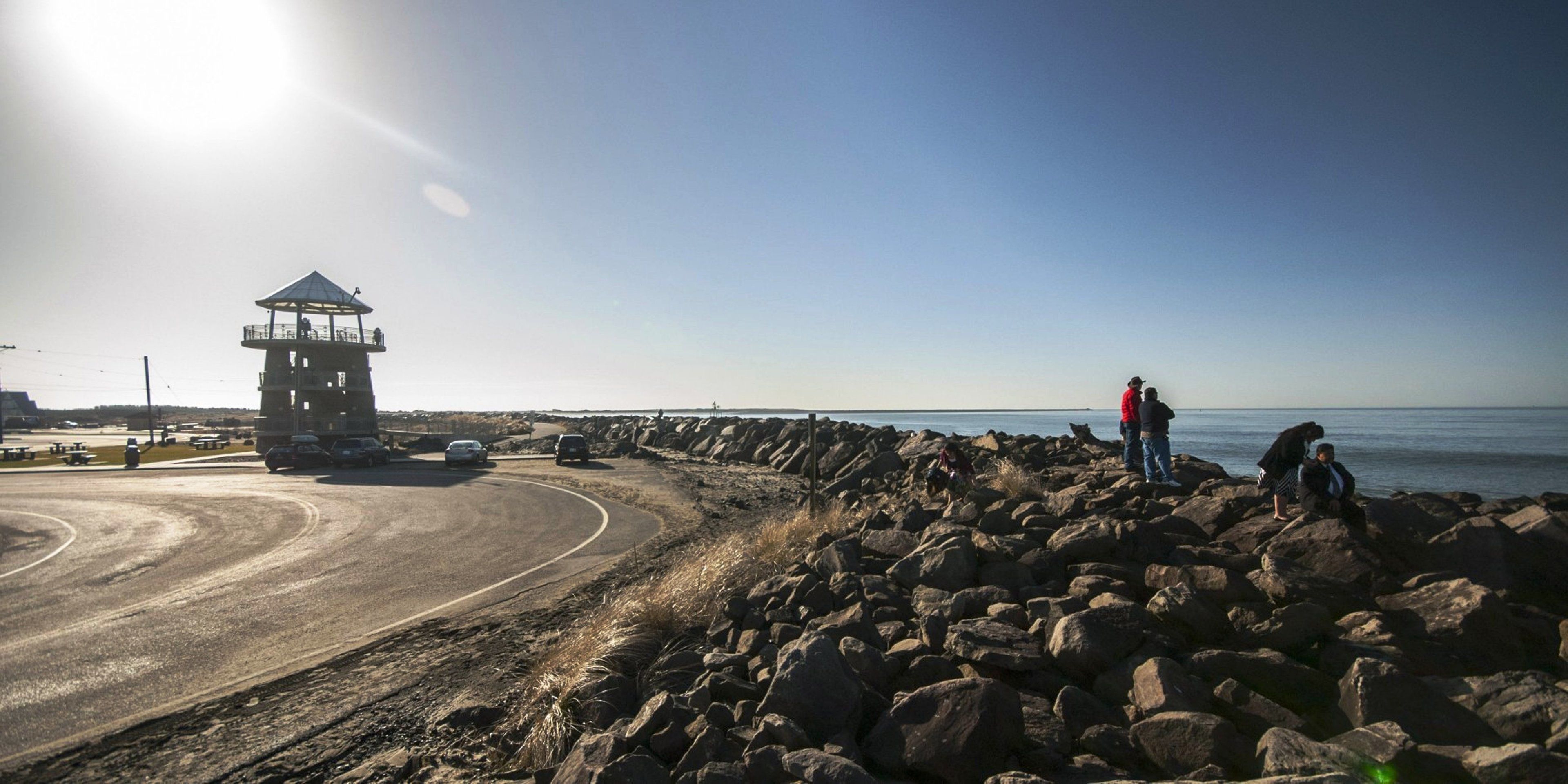 A coastal road with a guard tower, rocky shoreline, and a clear sky.
