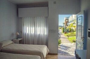 A hotel room with two beds, a nightstand with a lamp, a large window with curtains, and a view of a building and pathway outside.