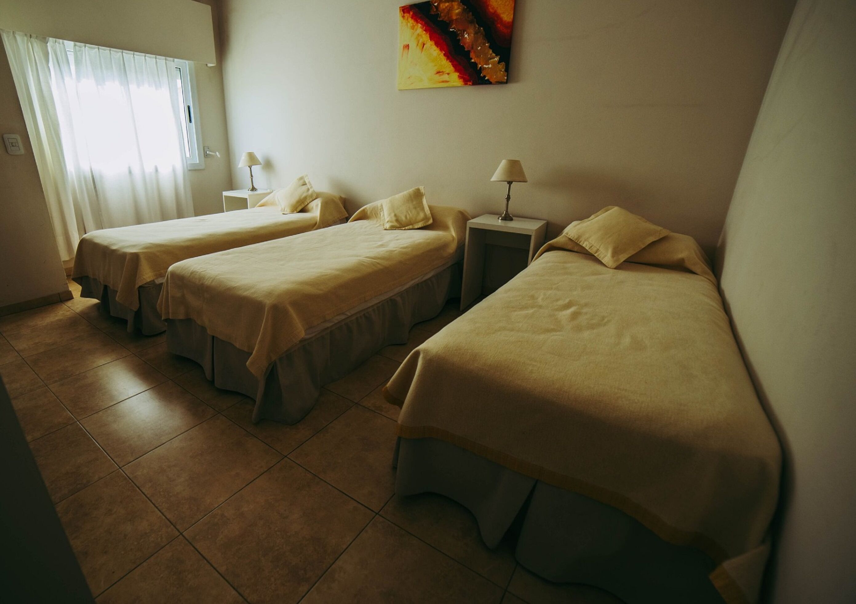 A hotel room with two single beds, a small table, a lamp, and a painting on the wall.