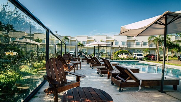 Ein Poolbereich mit Holzliegen und einem Tisch, mit Blick auf einen Pool und ein Gebäude mit mehreren Balkonen.