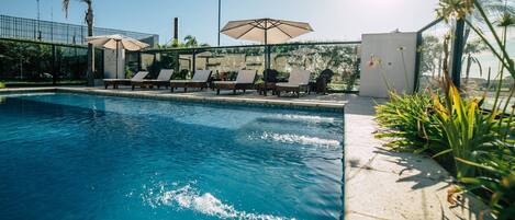 A swimming pool with lounge chairs and umbrellas, surrounded by greenery and a clear sky.