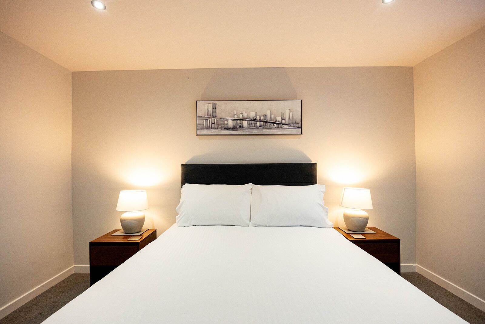 A hotel room with a large bed, two bedside tables with lamps, and a cityscape painting on the wall.