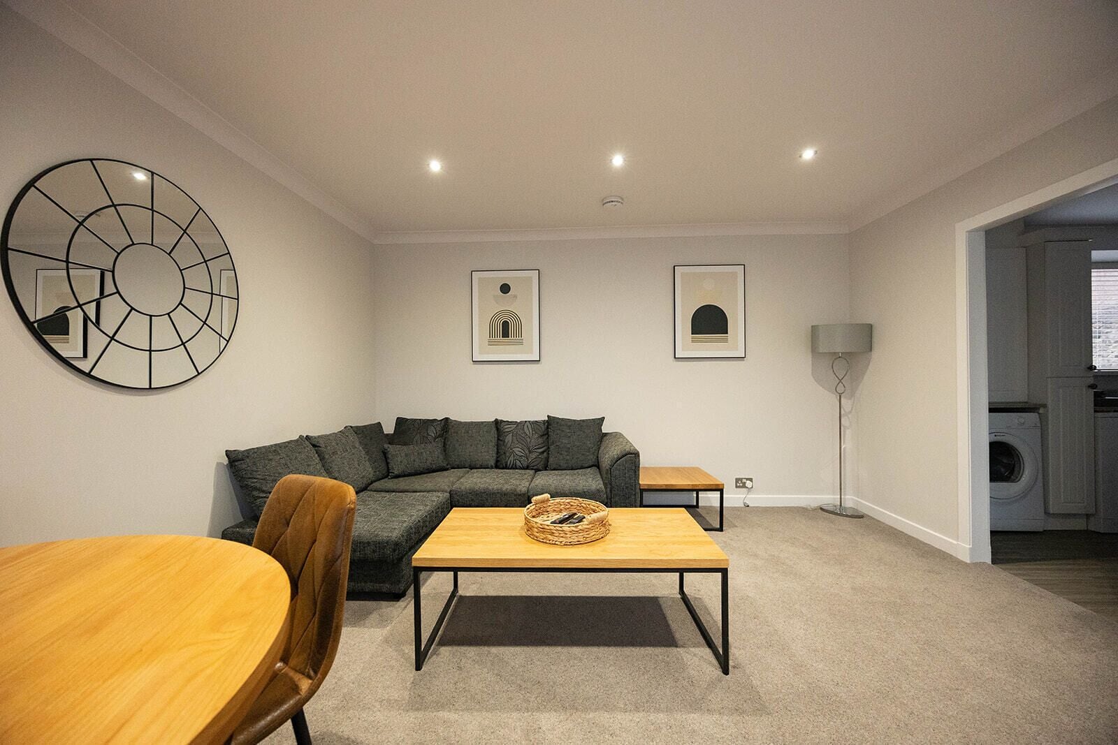 A modern living room with a sectional sofa, a wooden coffee table, and a dining table. There are two framed artworks on the wall and a floor lamp.