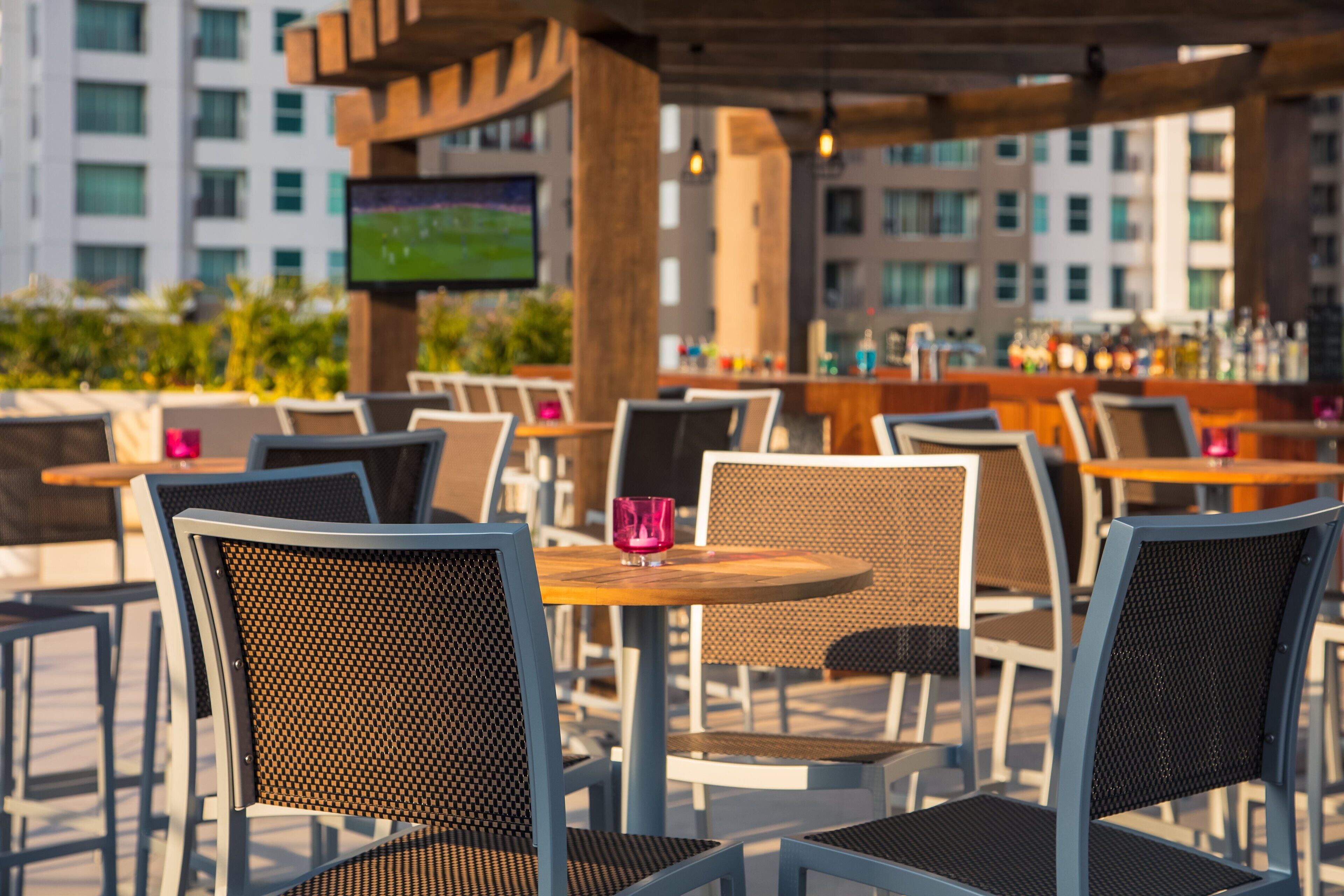 Outdoor seating with tables and chairs, a bar area, and a television screen in the background.