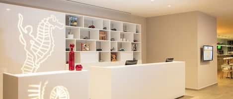A modern reception area with a white reception desk, a wall-mounted shelf with various items, and a digital display.