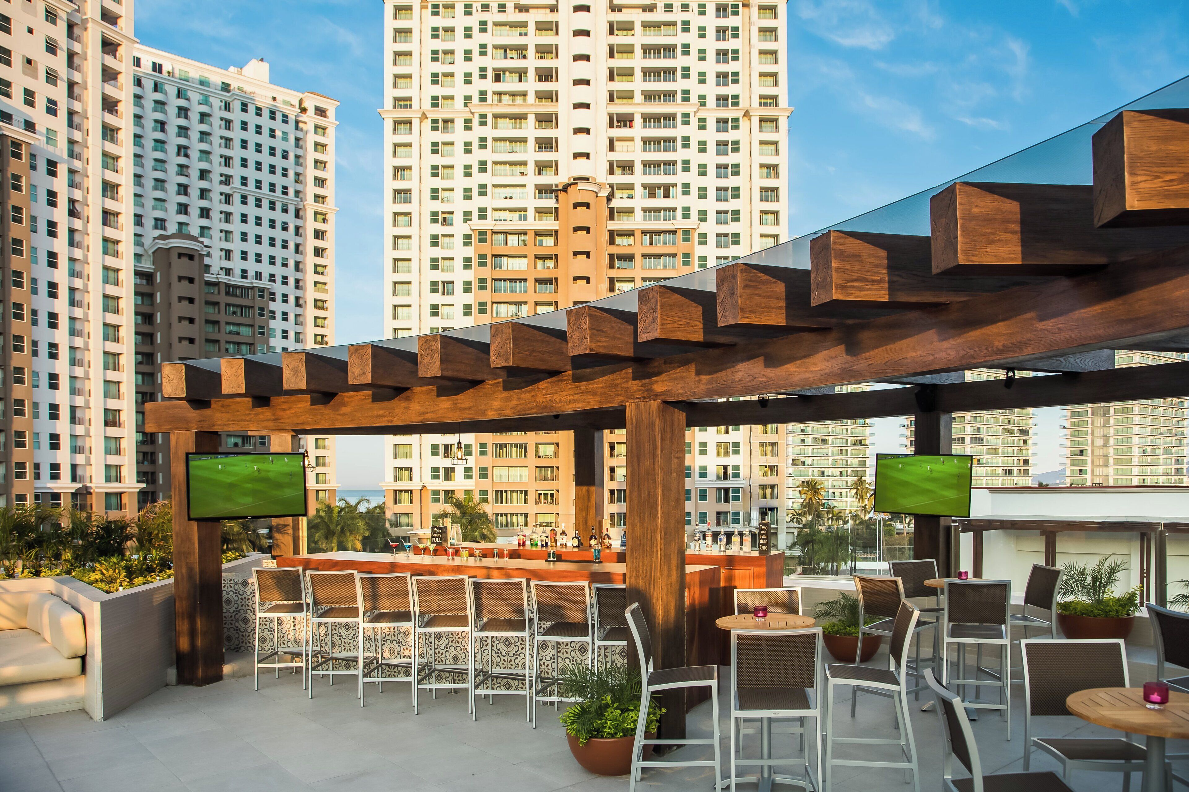 An outdoor bar area with seating, a TV screen, and a view of tall buildings.