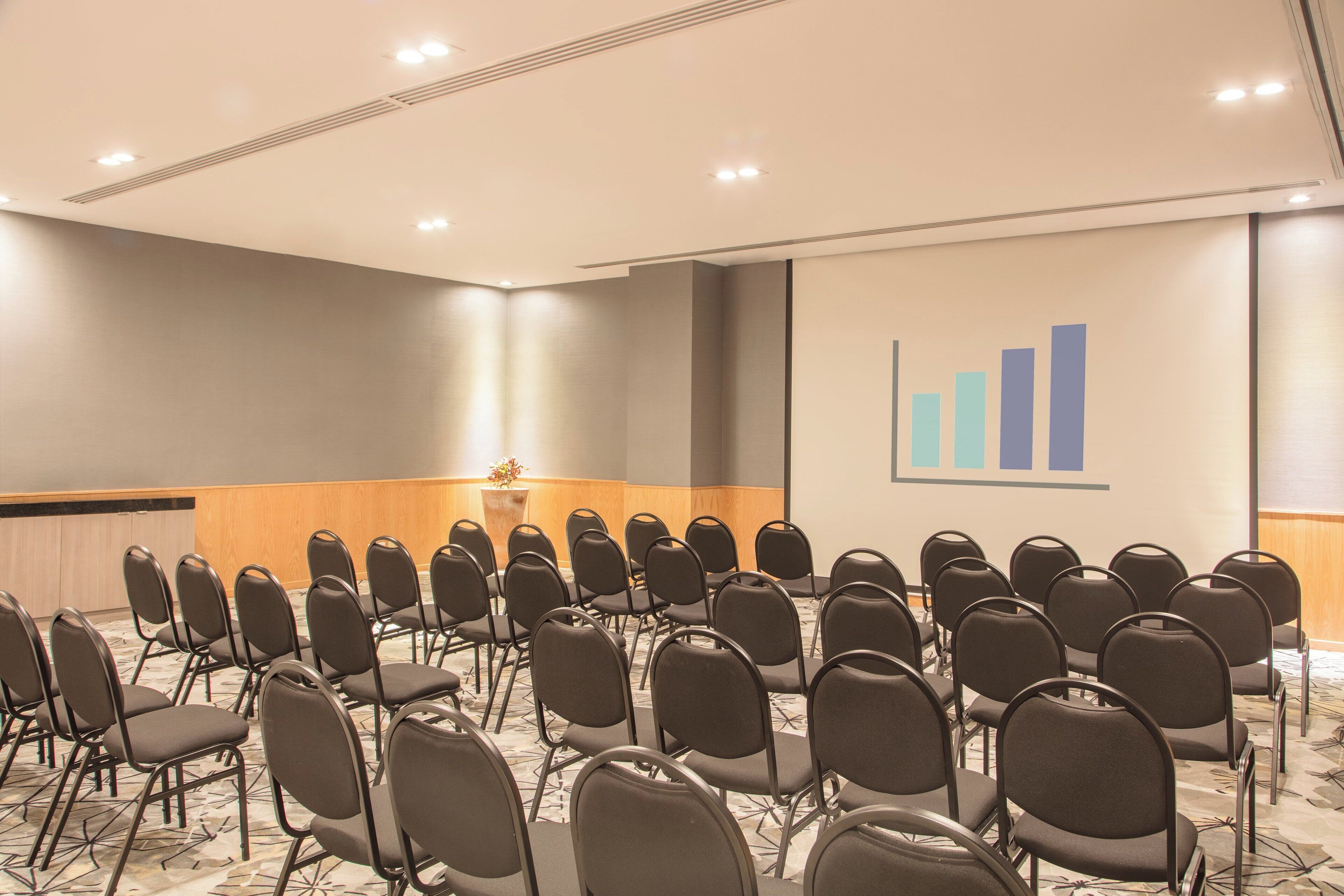 Un salón de conferencias con filas de sillas, un atril y una pantalla para presentaciones con un gráfico de barras.