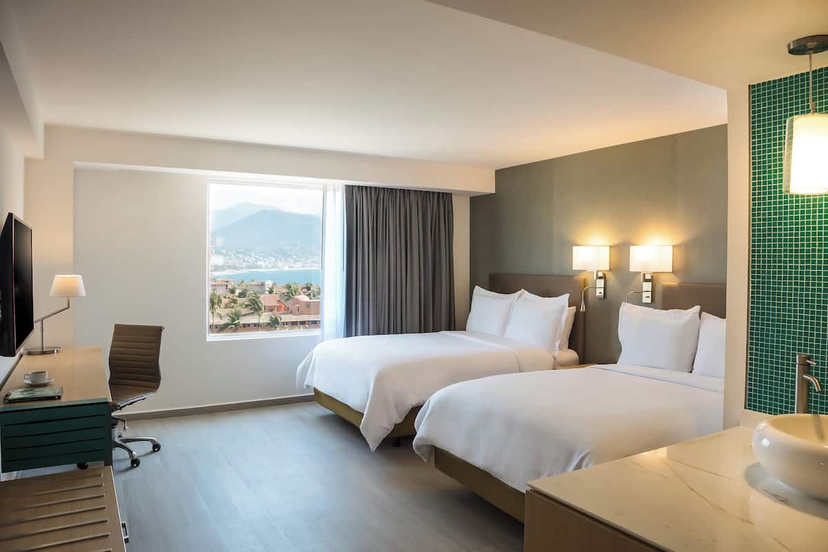 A hotel room with two beds, a desk with a chair, a TV, and a bathroom with a sink and mirror.