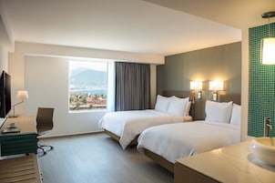A hotel room with two beds, a desk with a chair, a TV, and a bathroom with a sink and mirror.