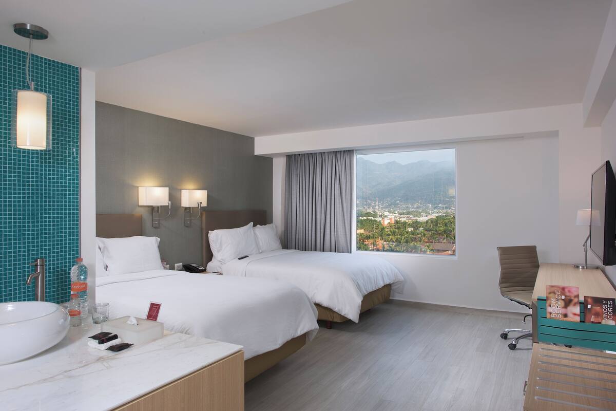 A hotel room with two beds, a large window with a view of mountains, a bathroom with a sink and mirror, and a desk with a chair.