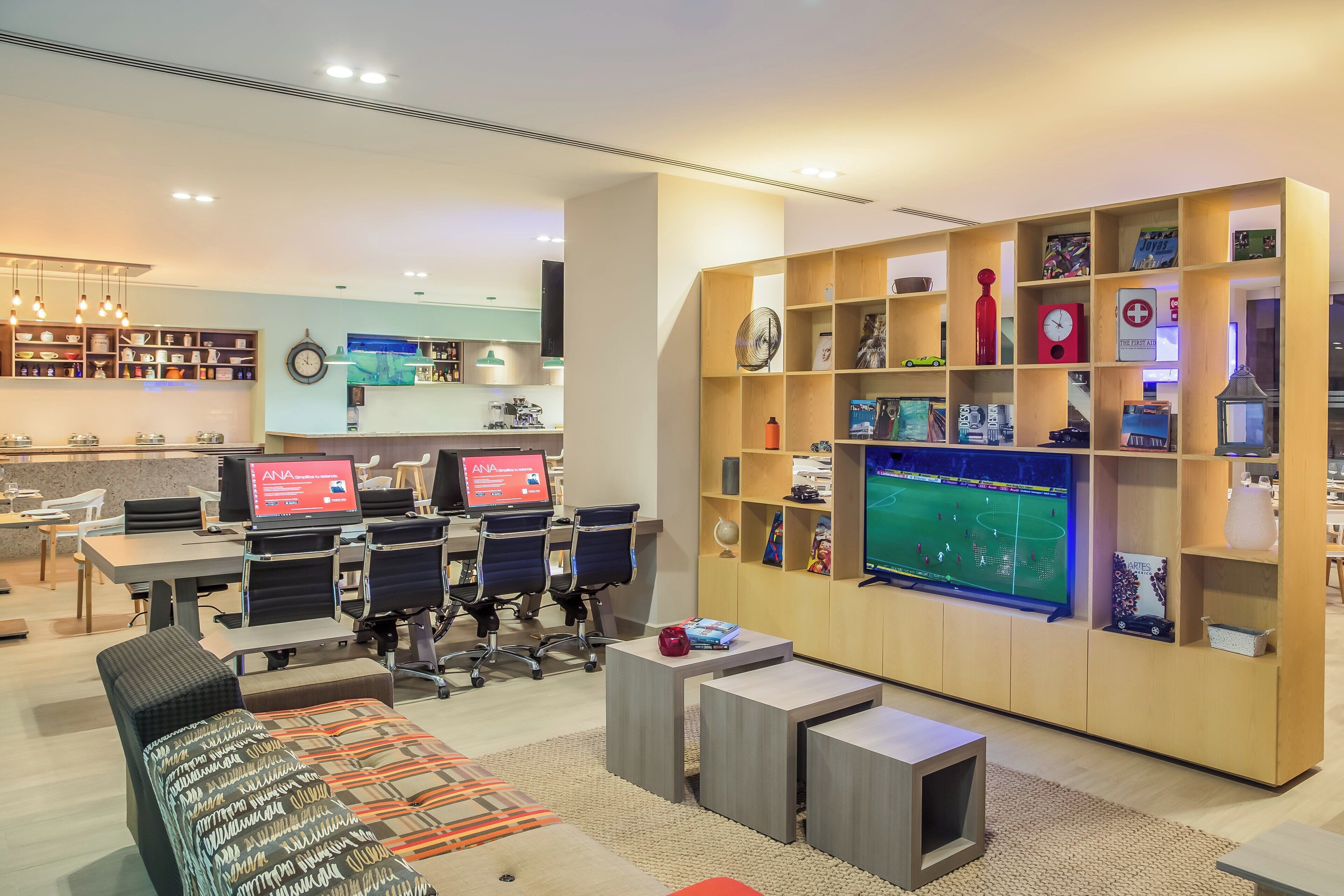 A modern hotel lobby with a reception desk, seating area, and a large TV screen displaying a sports game.