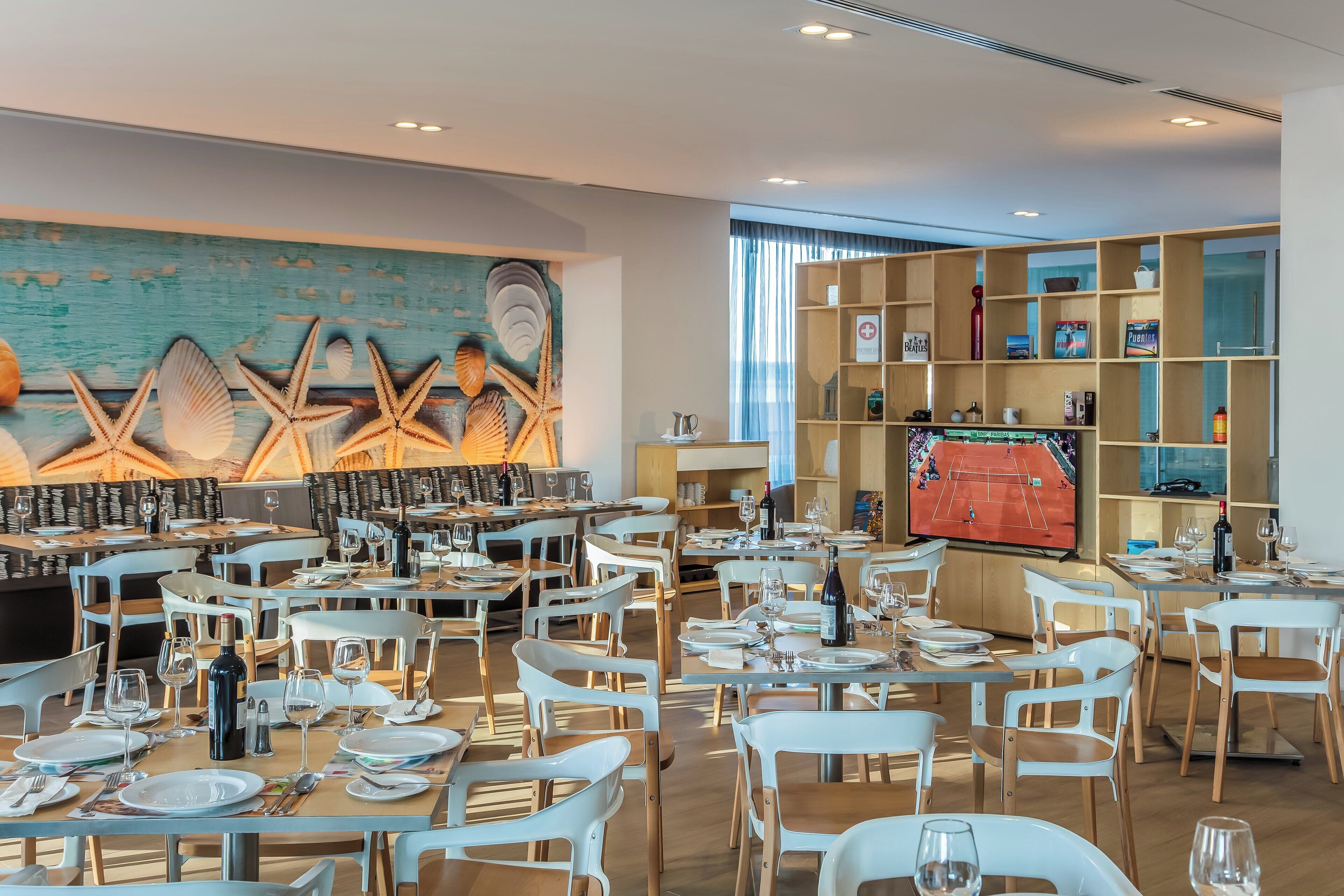 A restaurant with white chairs and tables, a mural of seashells, and a television displaying a tennis match.