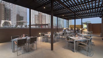 An outdoor dining area with tables and chairs, large umbrellas, and a view of high-rise buildings in the background.