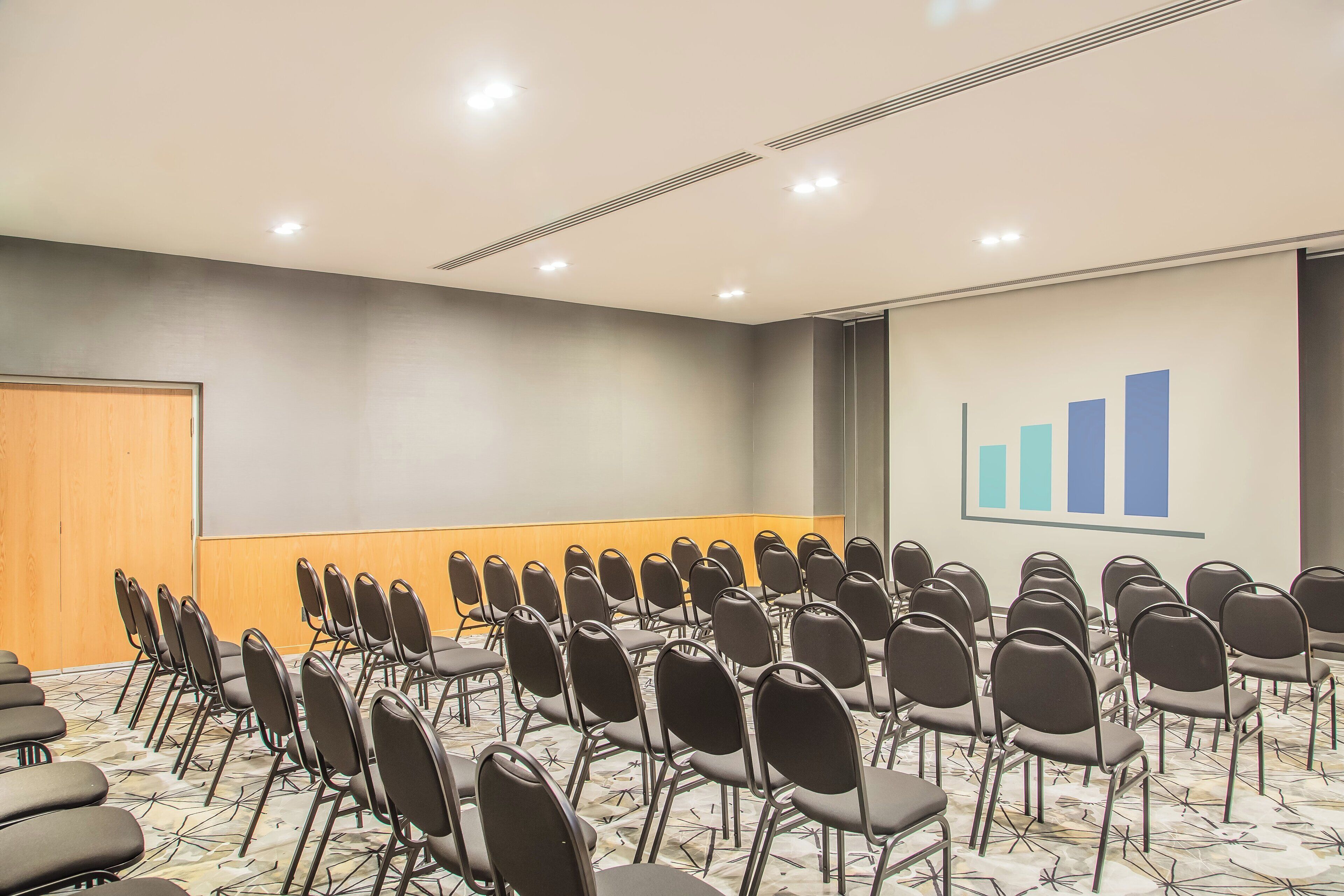 Un salón de conferencias con filas de sillas, una puerta de madera, una pizarra y un gráfico de barras en la pared.