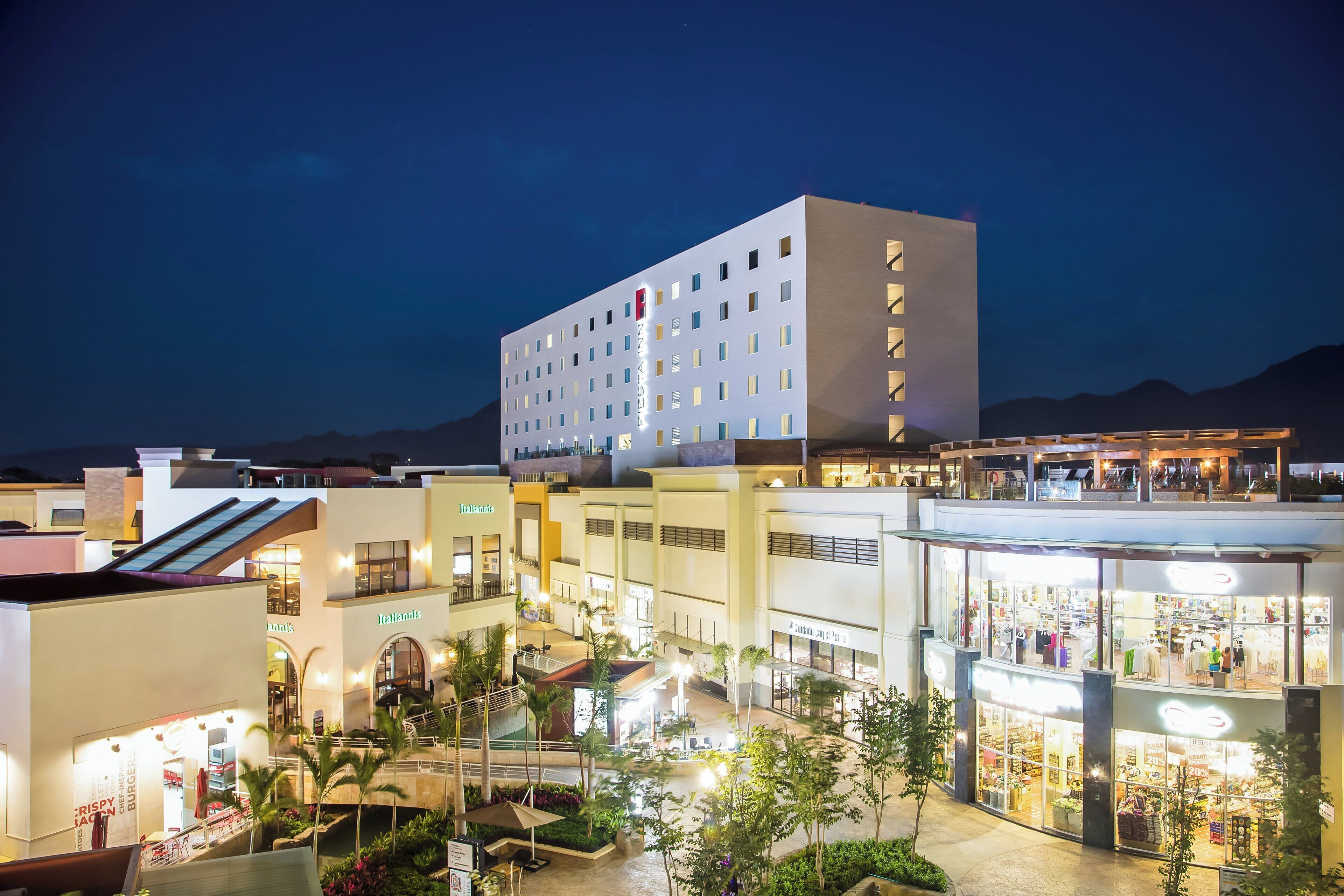 Un hotel de varios pisos con un diseño moderno, que cuenta con un patio central con plantas y mobiliario exterior.