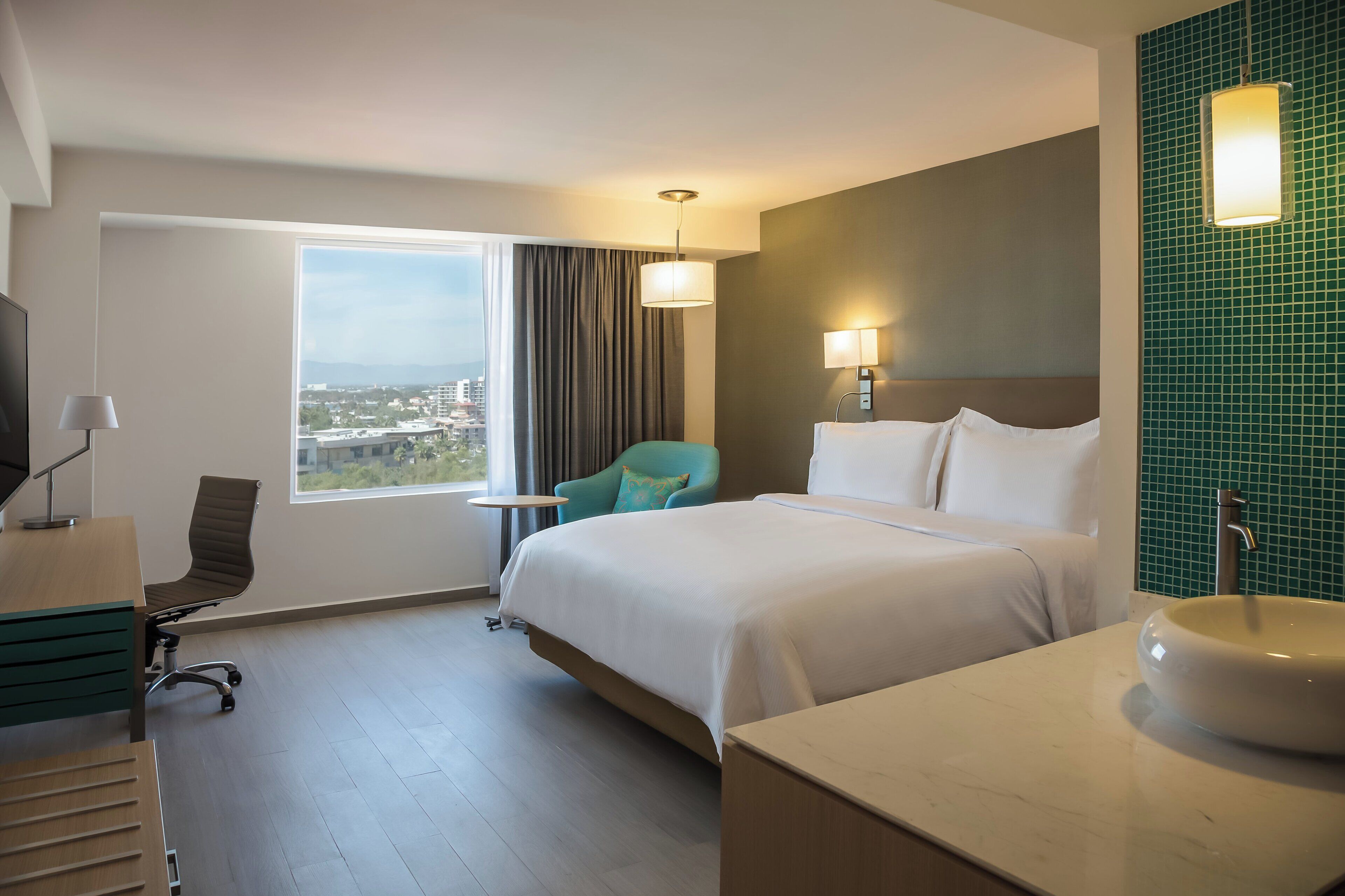 Une chambre d’hôtel avec un grand lit, un bureau avec une chaise, une petite table, une fenêtre donnant sur un beau paysage et une salle de bain avec un lavabo et un miroir.