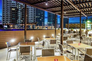 An outdoor dining area with tables and chairs, a pool, and a view of high-rise buildings at dusk.