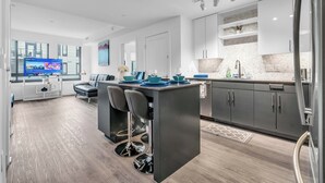 A modern kitchen with a central island, bar stools, and a dining area with a TV displaying a cooking program. - Fully Furnished Apartments near Nationals Park (Washington)