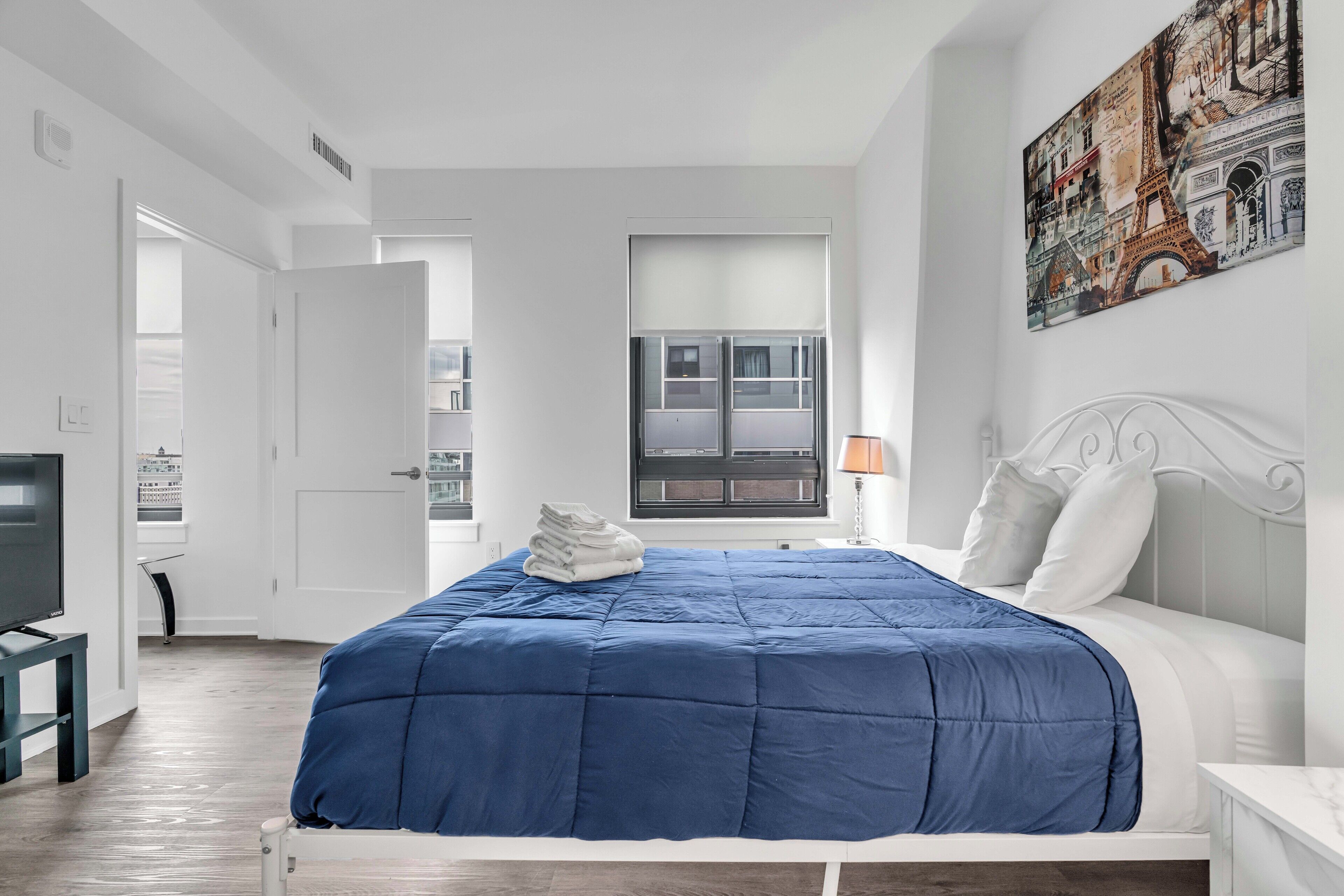 A bedroom with a bed, a painting of the Eiffel Tower, and a window with blinds.
