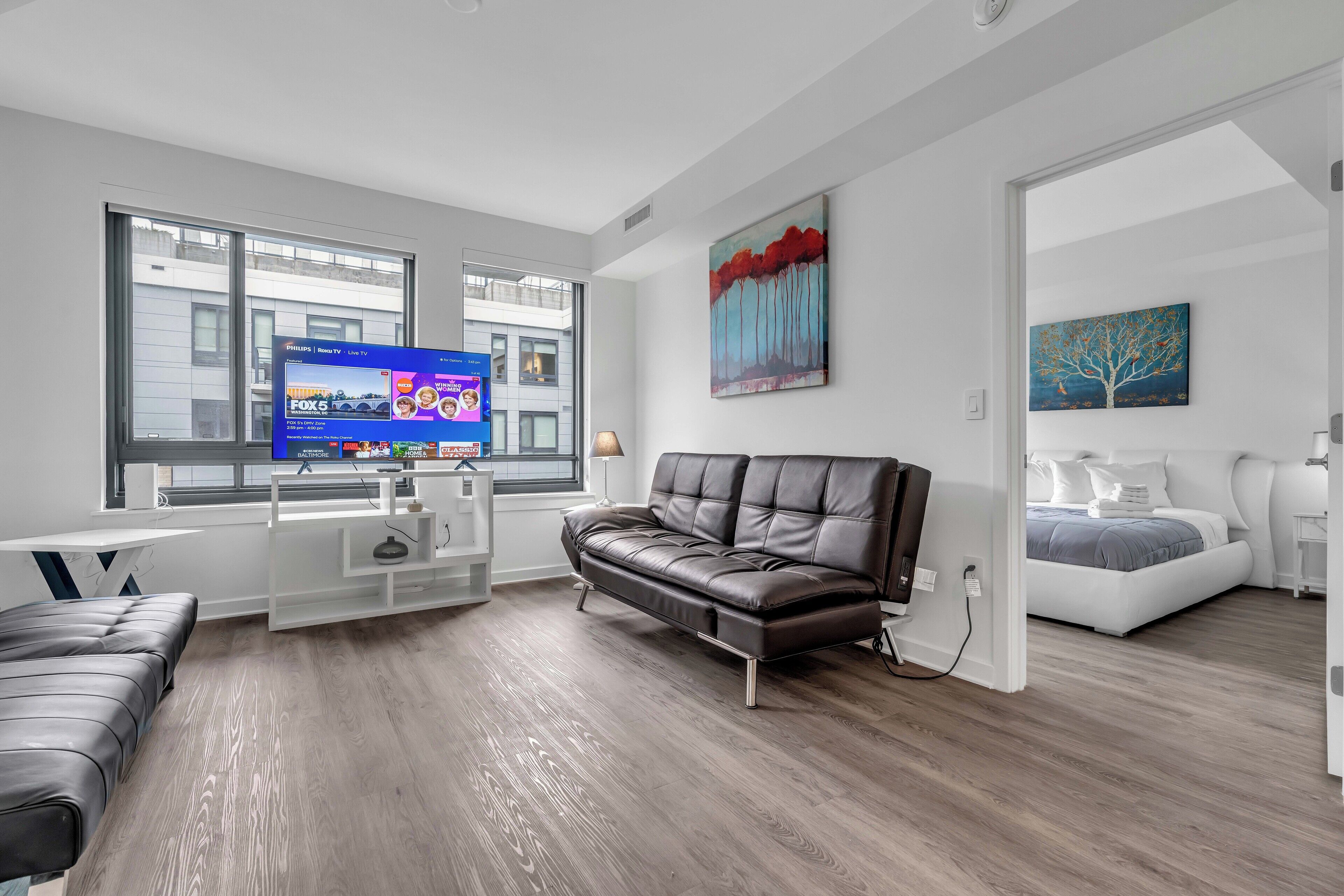 A modern living room with a leather sofa, a white entertainment unit, and a large window with a view of buildings.