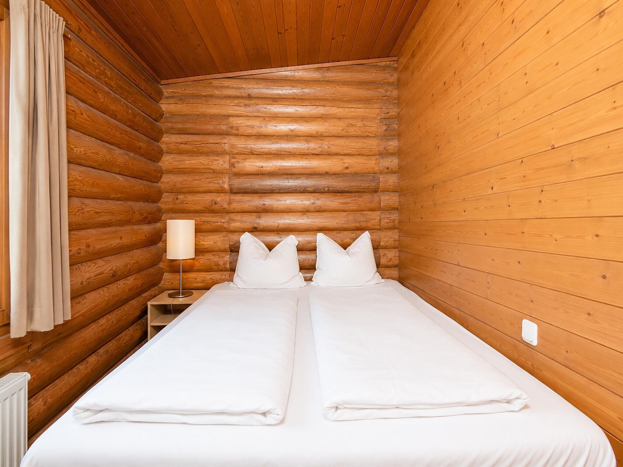 A log cabin room with two beds, a nightstand, a lamp, and a window with curtains.