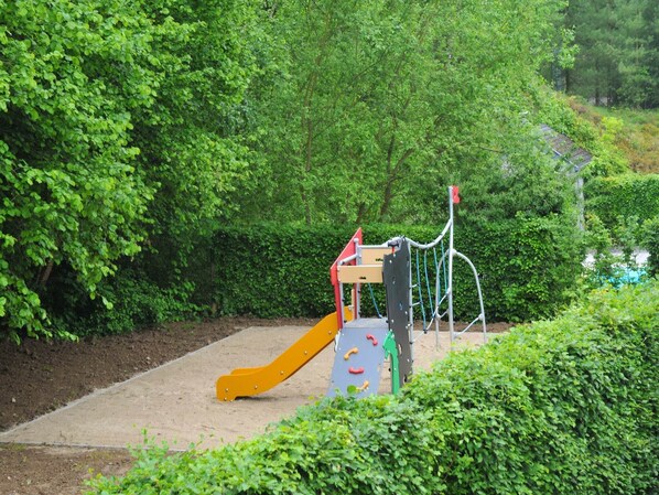 Un parque infantil con un tobogán y estructuras de escalada, rodeado de abundante vegetación.