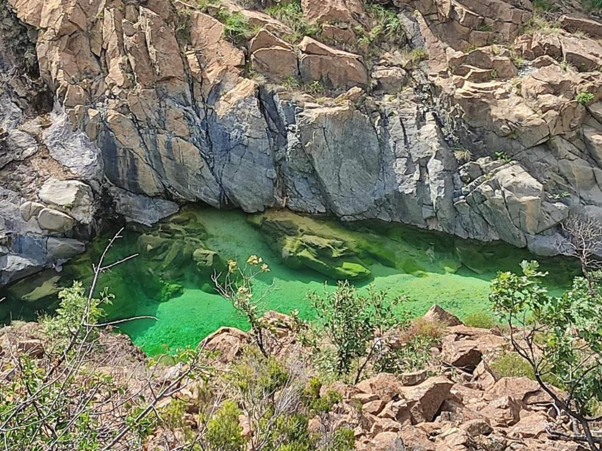 Eine felsige Landschaft mit einem klaren, grünen Pool, umgeben von zerklüftetem Gelände und spärlicher Vegetation.