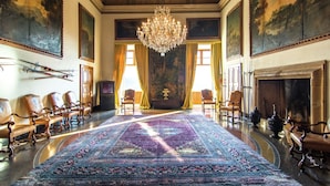 Een ruime kamer met een grote, versierde kroonluchter, een lang Perzisch tapijt en een open haard.