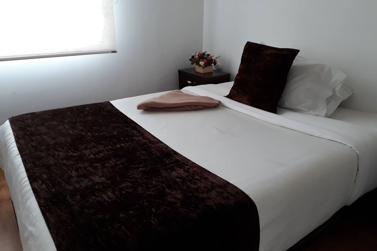 Ein ordentlich gemachtes Bett mit brauner Decke und weißen Laken, ein kleiner Nachttisch mit Blumen in einer Vase und ein Fenster mit Jalousien.