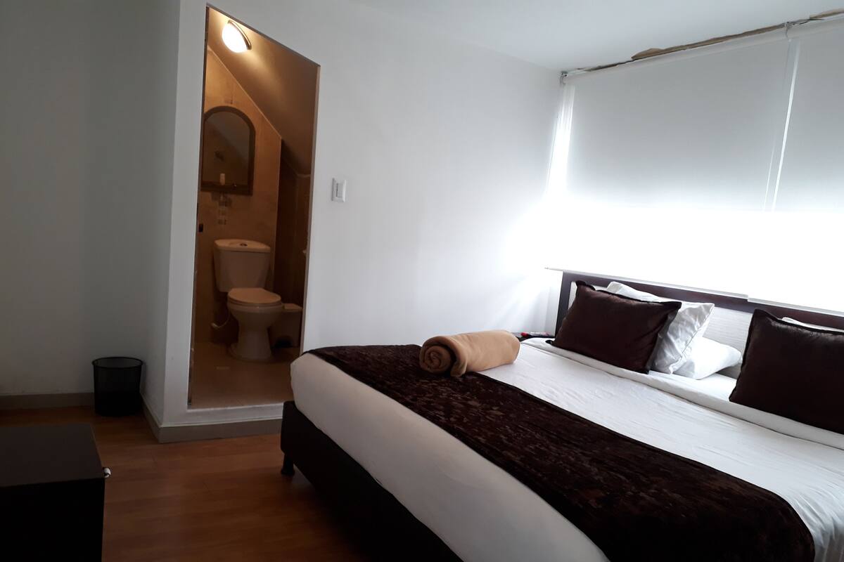 Ein Schlafzimmer mit einem Bett, einem Fenster, einer Tür zu einem Badezimmer und einer kleinen Mülltonne.