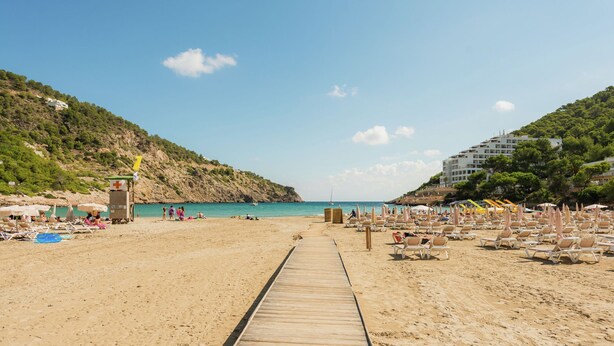 A beach with a wooden walkway leading to the water, beach chairs, umbrellas, and a lifeguard tower. - Villa in Cala Llonga near Beach & Ferry Port (Cala Llonga (santa Eulalia del Rio))