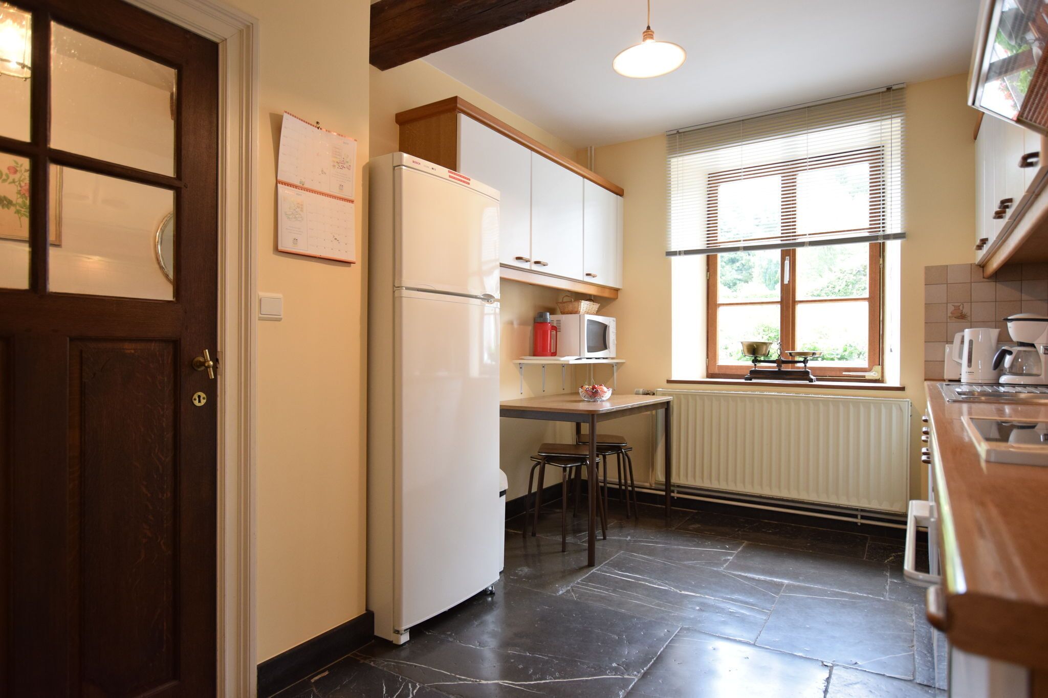 Een kleine keuken met een koelkast, magnetron en een raam met jaloezieën.