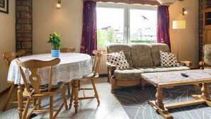 A cozy living room with a dining table, wooden chairs, a sofa with patterned cushions, and a potted plant.