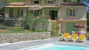 Une maison à deux étages avec une façade en pierre, des volets verts et un toit en tuiles. On y trouve une piscine avec des chaises longues jaunes et un mur en pierre au premier plan.