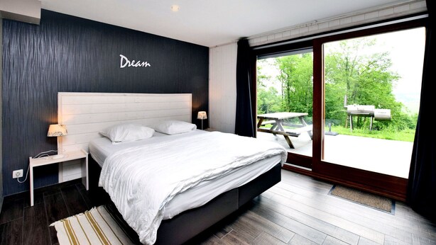 A modern bedroom with a large bed, a nightstand, a lamp, and a view of a garden through a sliding door. - Luxurious Chalet in Beauraing with Sauna (Beauraing)