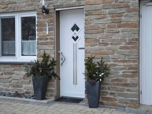 Un bâtiment en pierre avec une porte blanche, deux plantes en pot et une fenêtre avec un rideau.