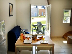 Un coin repas avec une table dressée pour deux personnes, un canapé et une vue sur un jardin par une porte ouverte.